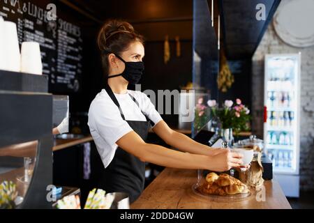 Barista femminile in maschera protettiva che dà caffè durante il lavoro al caffè Foto Stock