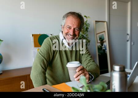 Uomo maturo sorridente che tiene una tazza di caffè usa e getta seduto a casa Foto Stock