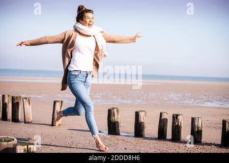 Felice giovane donna con braccia allungate che corrono tra pali di legno in spiaggia durante il giorno di sole Foto Stock