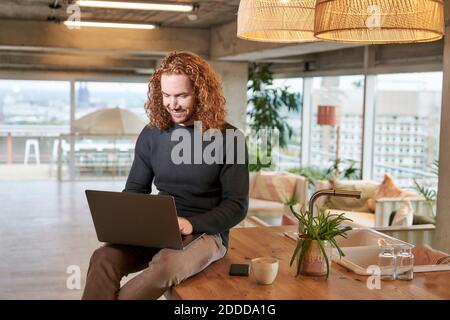 Sorridente uomo rosso che usa un computer portatile seduto sul tavolo a casa Foto Stock