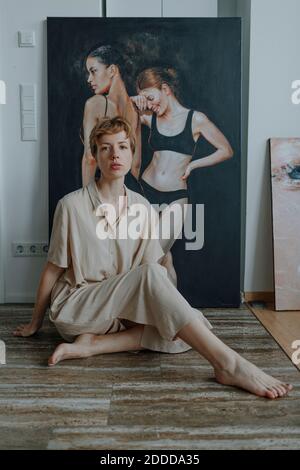 Fidato artista femminile di metà adulto seduto sul pavimento di legno contro pittura Foto Stock