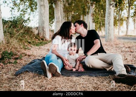 I genitori baciano mentre si siedono con il bambino bot sulla coperta all'aperto Foto Stock