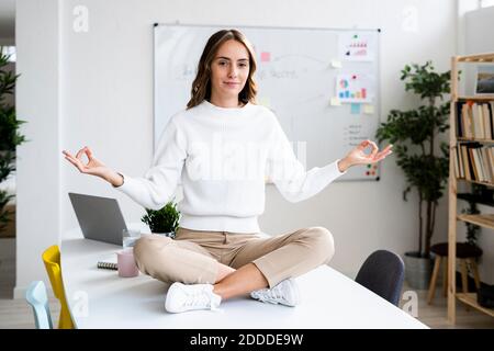 Giovane donna d'affari che fa yoga mentre si siede sul tavolo in ufficio Foto Stock