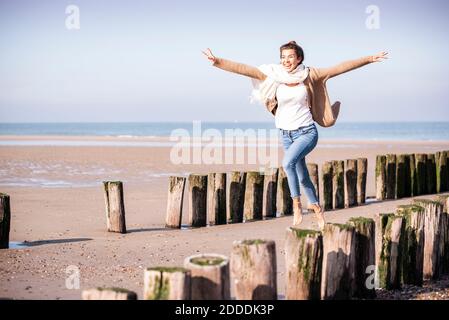 Giovane donna allegra con braccia allungate che corrono tra pali di legno in spiaggia durante il giorno di sole Foto Stock