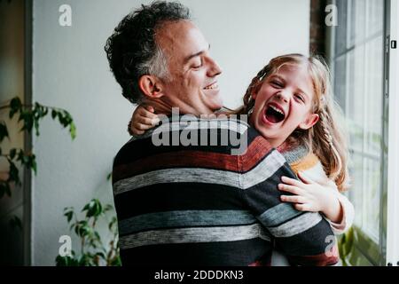 Sorridente padre che porta la figlia giocosa mentre si sta in piedi vicino alla finestra a. casa Foto Stock