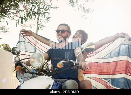 Donna sorridente con le braccia allungate tenendo bandiera nazionale mentre si siede dietro partner sulla moto durante l'estate Foto Stock