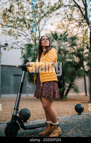 Donna premurosa con scooter elettrico a spinta in strada durante l'autunno Foto Stock