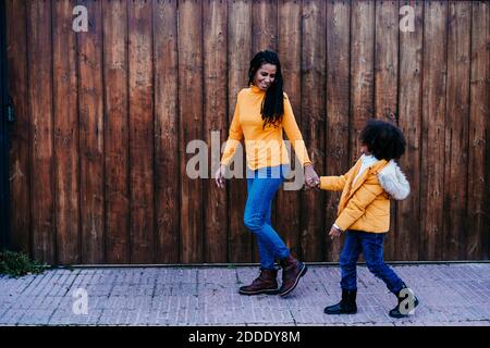 Madre che tiene la mano della figlia mentre cammina contro il muro di legno Foto Stock