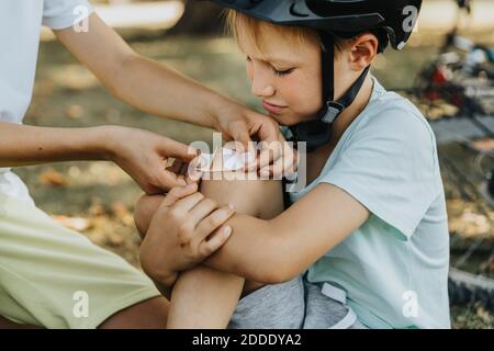 Ragazzo che mette il bendaggio sul ginocchio del fratello più giovane seduto in pubblico parcheggio Foto Stock