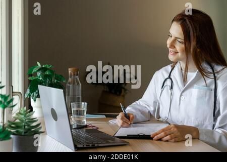 Giovane dottoressa che scrive rapporto medico durante la consultazione online da ufficio domestico Foto Stock