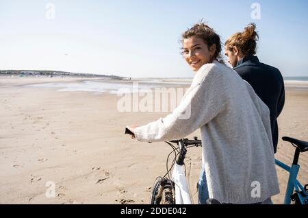 Sorridente bella donna che cammina in bicicletta da ragazzo mentre si guarda oltre la spalla in spiaggia contro il cielo limpido in giornata di sole Foto Stock