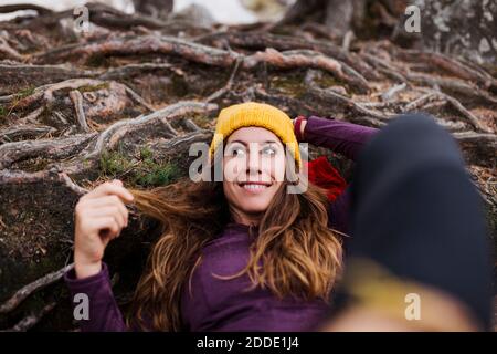 Donna sorridente che guarda via mentre si sdraia sulle radici degli alberi nella foresta a la Pedriza, Madrid, Spagna Foto Stock