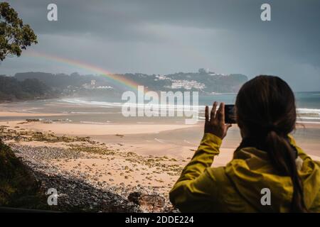 Vista posteriore della donna in impermeabile fotografando l'arcobaleno sulla spiaggia Foto Stock