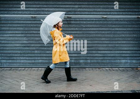 Ragazza che indossa impermeabile e scarpone da salto che tiene l'ombrello mentre cammina sul marciapiede Foto Stock