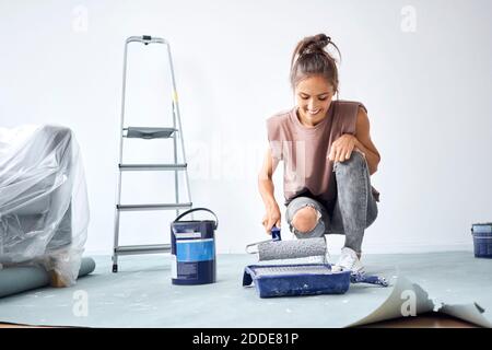 Donna sorridente che tiene il rullo di vernice mentre accovacciandosi a casa Foto Stock