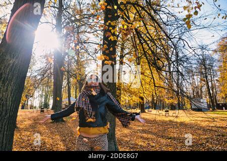 Donna felice in piedi con le braccia allungate sotto caduta autunno asciutto foglie mentre contro gli alberi al parco Foto Stock