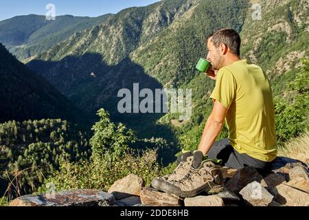 Uomo medio adulto che beve caffè mentre si siede sulla pietra dentro foresta durante il giorno di sole Foto Stock
