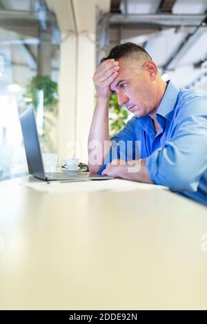 Un imprenditore maschile angosciato con la mano sulla fronte fissando il computer portatile mentre si è seduti nella caffetteria Foto Stock