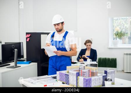 Giovane architetto maschile in un cappello bianco e tute che lavorano in ufficio. Foto Stock