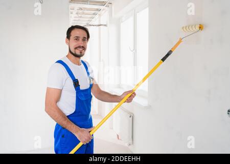 Colpo medio, operaio generale in abiti da lavoro blu dipinge parete in appartamento Foto Stock