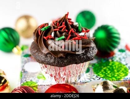 Una Cupcake di cioccolato di Natale si siede su un regalo di foglio avvolto con ornamenti di Natale sparsi intorno. Sfondo bianco. Foto Stock