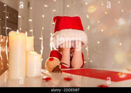 Bambina felice in un vestito rosso e cappello scrive una lettera a Babbo Natale. Il bambino fa un desiderio per Natale. Il bambino non sa cosa fare una g Foto Stock