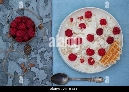 Ricotta il formaggio in un piatto con yogurt, cosparso di lamponi e muesli, blu su un asciugamano, cucchiaio di metallo e noci. Foto Stock