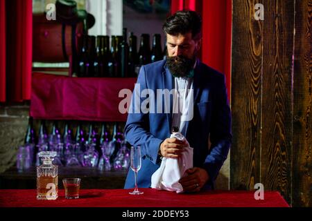 Bel barman con bearded e barba lunga e baffi con serio cocktail alcolico fatto sul viso in pelle scamosciata vintage su sfondo viola rosso bar. Foto Stock