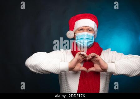 Un uomo in un cappellino e maschera di Natale fa un gesto cardiaco con le dita.incontro concettuale, congratulazioni per il nuovo anno. Foto di alta qualità Foto Stock