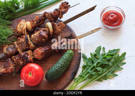 Maiale alla griglia su spiedini di metallo arrostito su una griglia con rucola fresca, prezzemolo, aneto, erba cipollina su trivella di legno con pomodoro, cetriolo e pomodoro Foto Stock