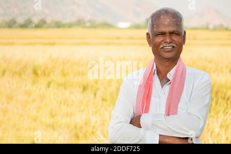 Ritratto di sicuro sorridente agricoltore indiano con le braccia incrociate in piedi di fronte al terreno agricolo guardando fotocamera con copia spazio.