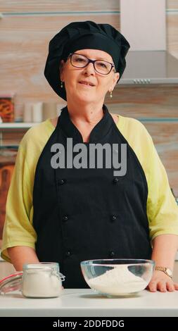Donna anziana felice che indossa bonete guardando la camerea in sala da pranzo a casa. Vecchio panettiere pensionato con uniforme da cucina preparazione ingredienti da forno a tavola pronti per cucinare pane, torte e pasta fatti in casa. Foto Stock