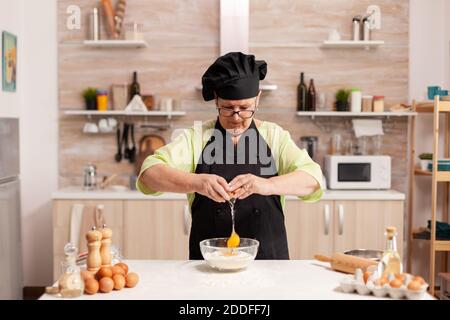 Donna anziana che cricchiola le uova sulla farina di grano nella cucina domestica. Cuoco di pasta anziano che coglie l'uovo sulla ciotola di vetro per la ricetta della torta in cucina, mescolando a mano, impastando Foto Stock