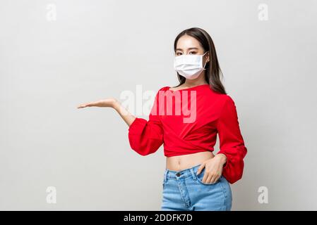 Donna asiatica indossando maschera medica e aprire la mano a vuoto spazio da parte su sfondo grigio chiaro studio Foto Stock