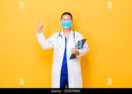 Giovane donna asiatica medico indossando maschera medica che punta la mano in su per svuotare lo spazio isolato su sfondo giallo Foto Stock