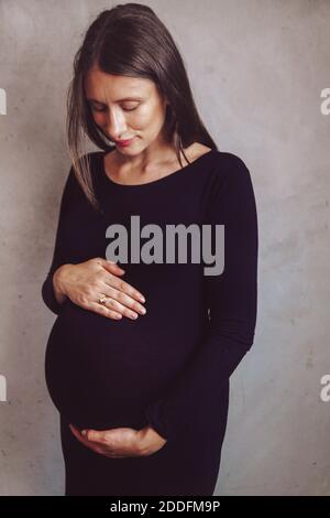 Ritratto di una giovane donna incinta dai capelli scuri in un abito nero su sfondo neutro. Urto di gravidanza. Concetto femminile di madre attesa. Foto Stock