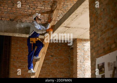 Giovane costruttore maschile in tute blu e elmetto che guarda messo a fuoco, salendo la scala mentre si lavora in cantiere. Costruzione, persone, concetto di professione. Vista laterale Foto Stock