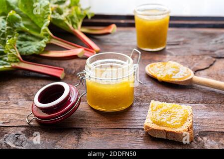 Vasetti di vetro di marmellata di rabarbaro fatta in casa, un toast con marmellata e gambi di rabarbaro fresco su un tavolo di legno scuro vecchio vicino alla finestra, sana colazione biologica Foto Stock