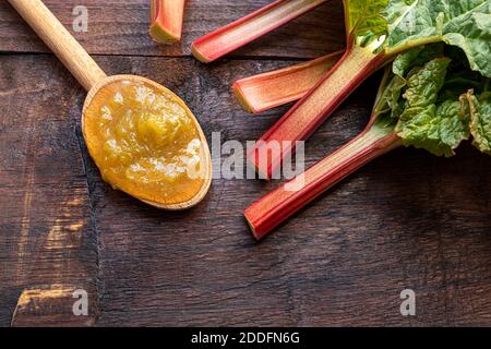 Un cucchiaio di legno con marmellata di rabarbaro fatta in casa e gambi di rabarbaro oraganico fresco su di un vecchio sfondo di legno scuro, concetto di dessert sano Foto Stock