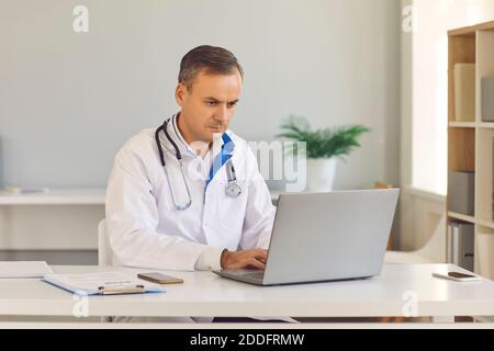 Medico maschile concentrato seduto in ufficio di fronte al computer portatile e rivedere i risultati dei test per la malattia Foto Stock