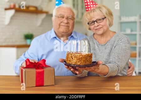 Sorridente coppia matura in caps vacanza seduta, tenendo torta di compleanno con candele e confezione regalo Foto Stock