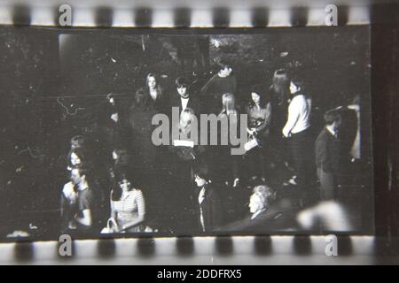 Bella fotografia in bianco e nero degli anni '70 di una folla di persone che aspettano in fila per un giro di carnevale al parco divertimenti locale in serata. Foto Stock