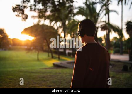 Ragazzo caucasico biondo che ascolta la musica con le cuffie wireless al tramonto, indossando un pullover rosso. Concetto di tecnologia. Musica per strada. Foto Stock