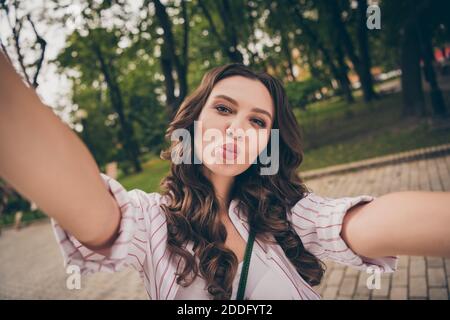 Ritratto fotografico di bella ragazza giovane con lunghi capelli ondulati presa del selfie che invia il bacio dell'aria con i labbri della groppa pouted dentro parco cittadino Foto Stock