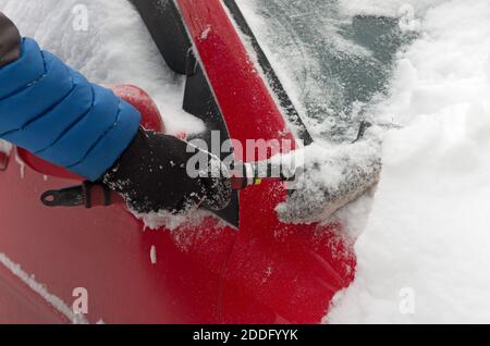 L'auto rossa viene pulita dalla neve con una spazzola accesa un closeup nuvoloso giorno d'inverno Foto Stock