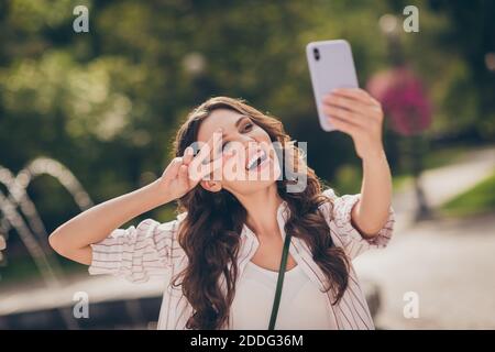 Foto di divertente ridere lunghi capelli castani ricci giovane donna fare selfie show v-sign godere weekend in park wear a strisce camicia esterna Foto Stock