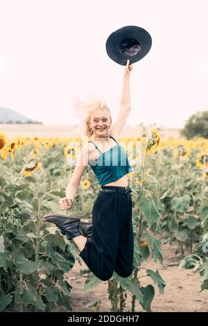 Corpo pieno di felice spensierata giovane bionda femmina in casual vestito con cappello in mano che si diverte e salta in alto mentre godendo la giornata estiva in girasole f Foto Stock