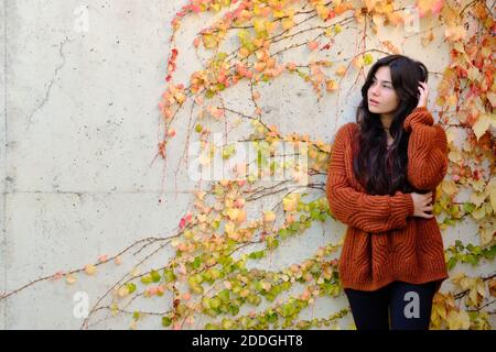 Bella bruna giovane con capelli lunghi in maglia calda in piedi vicino muro di pietra con coloratissima vegetazione autunnale e guardando lontano Foto Stock