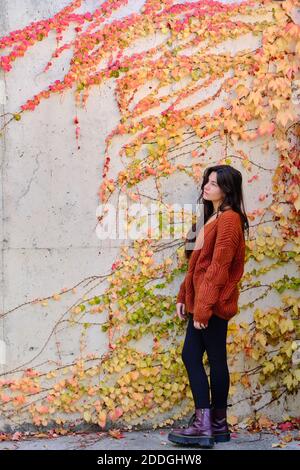 Bella bruna giovane con capelli lunghi in maglia calda in piedi vicino muro di pietra con coloratissima vegetazione autunnale e guardando lontano Foto Stock