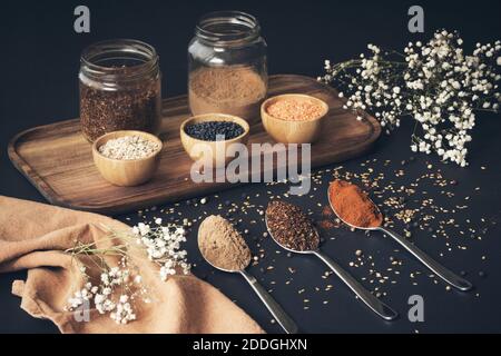 Vista dall'alto di cereali assortiti in vasi e spezie aromatiche in cucchiai disposti su vassoio di legno su tavolo nero Foto Stock